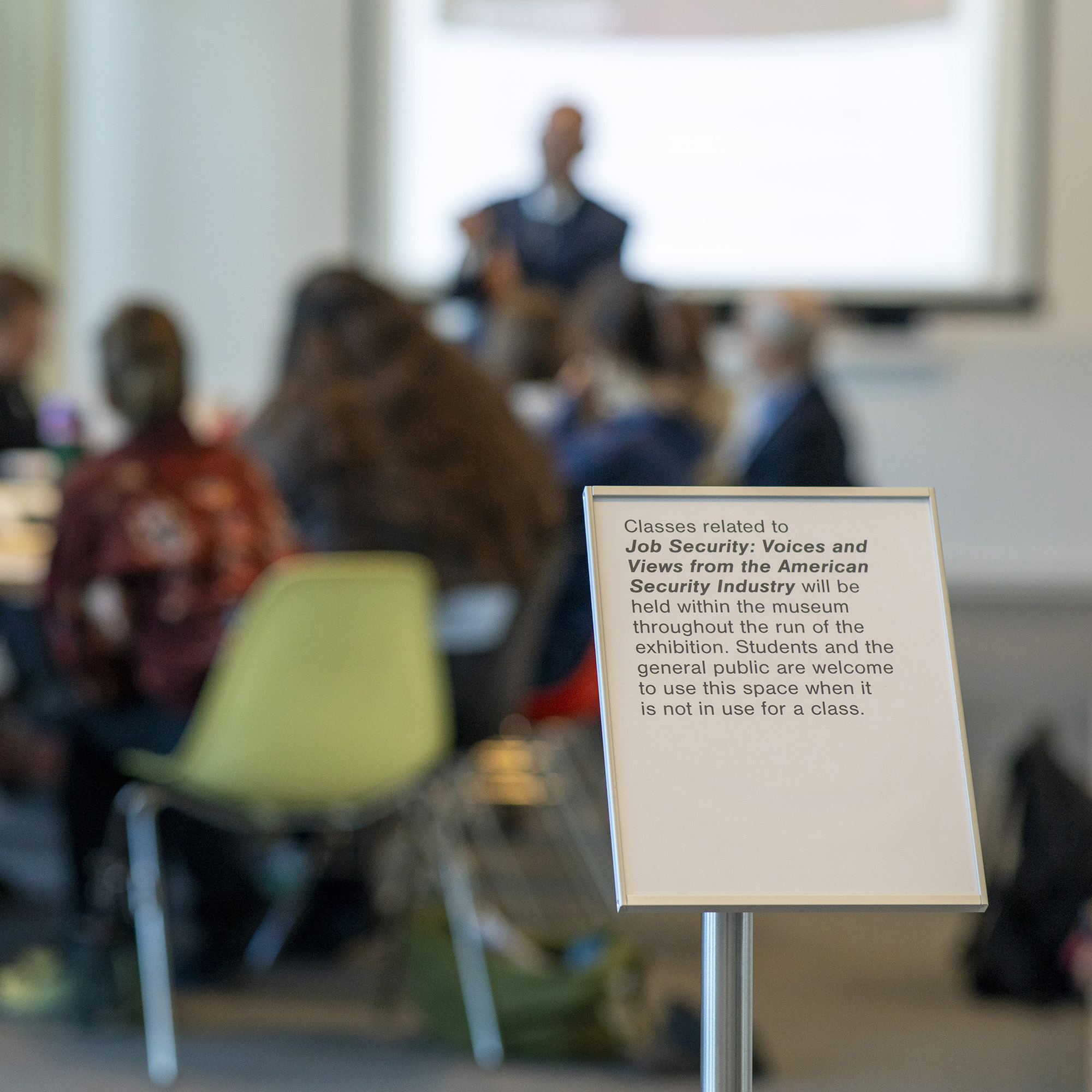 Shallow focus photograph of a presenter in front of a class with an aluminum sign in the foreground. Sign reads: Classes related to Job Security: Voices and Views from the American Security Industry will be held within the museum throughout the run of the exhibition. Students and the general public are welcome to use this space when it is not in use for a class.